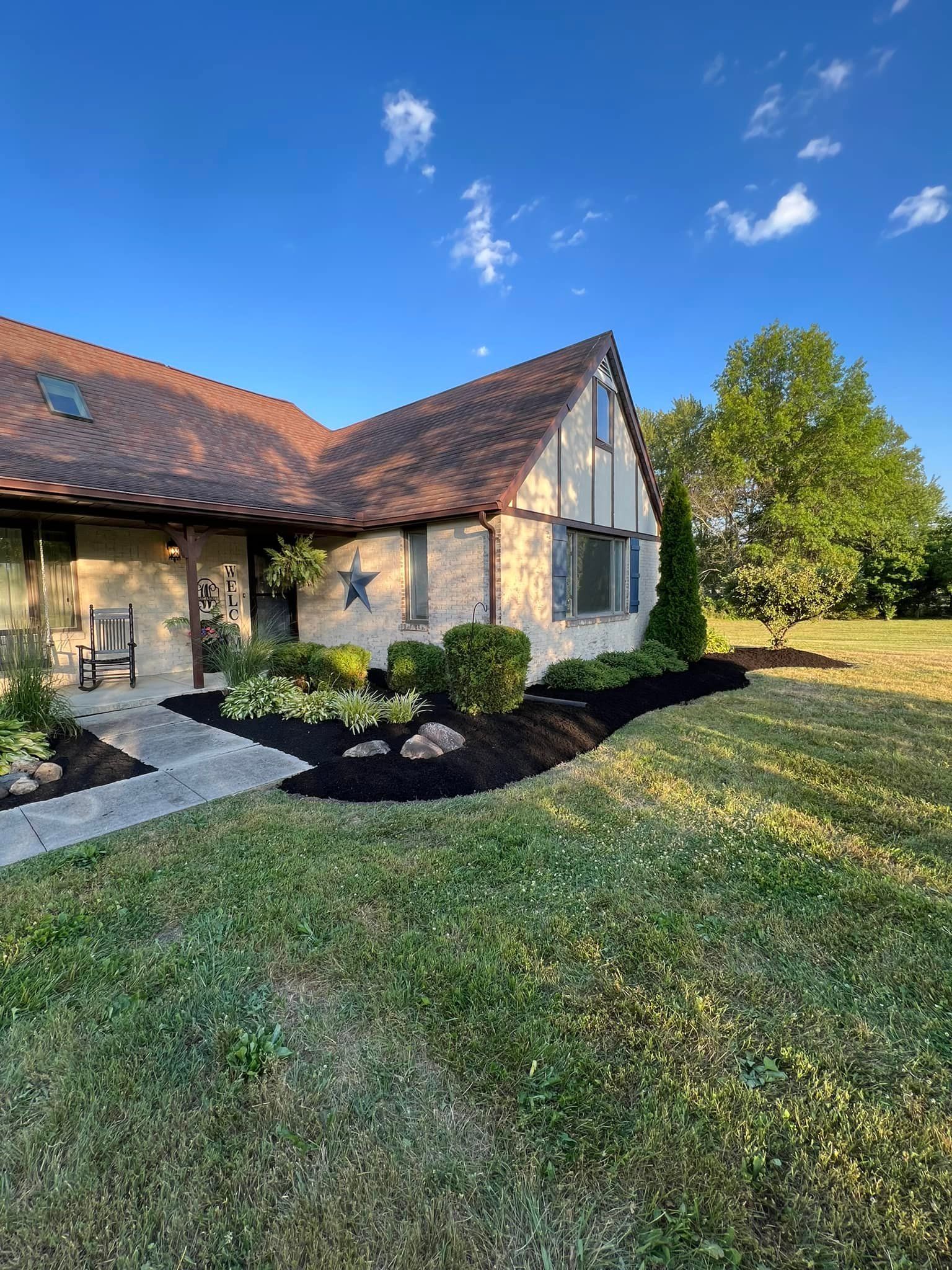 A brick house with a brown roof, black mulch, and a lush green lawn under a blue sky.