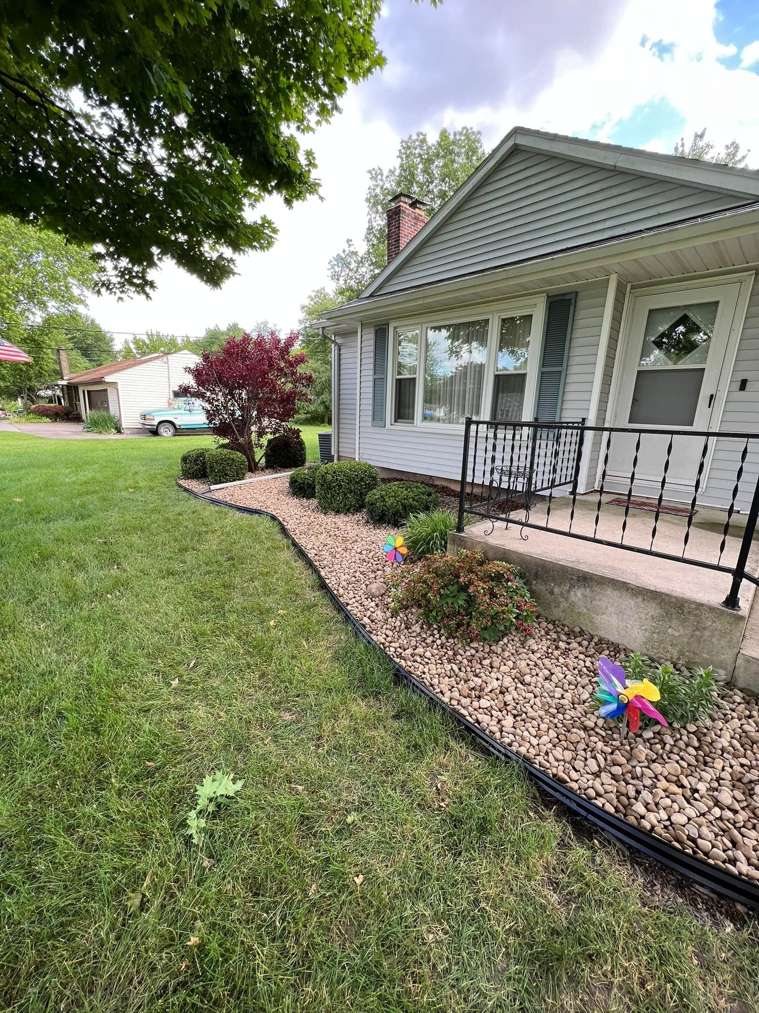 A house with a front yard flower bed. The yard has green grass, tan rocks, and colorful pinwheels.