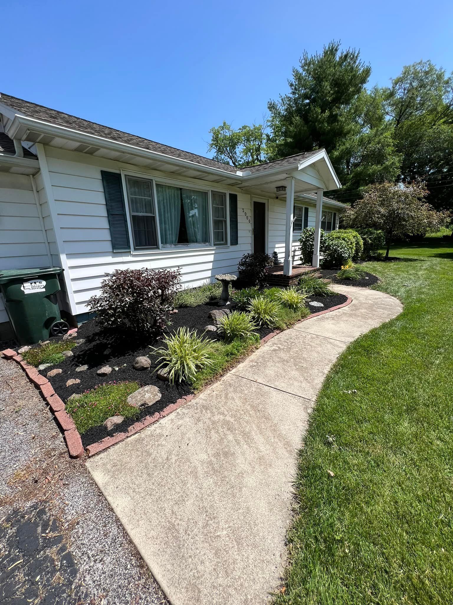 White house with a concrete path, flowerbeds, and green lawn on a sunny day.