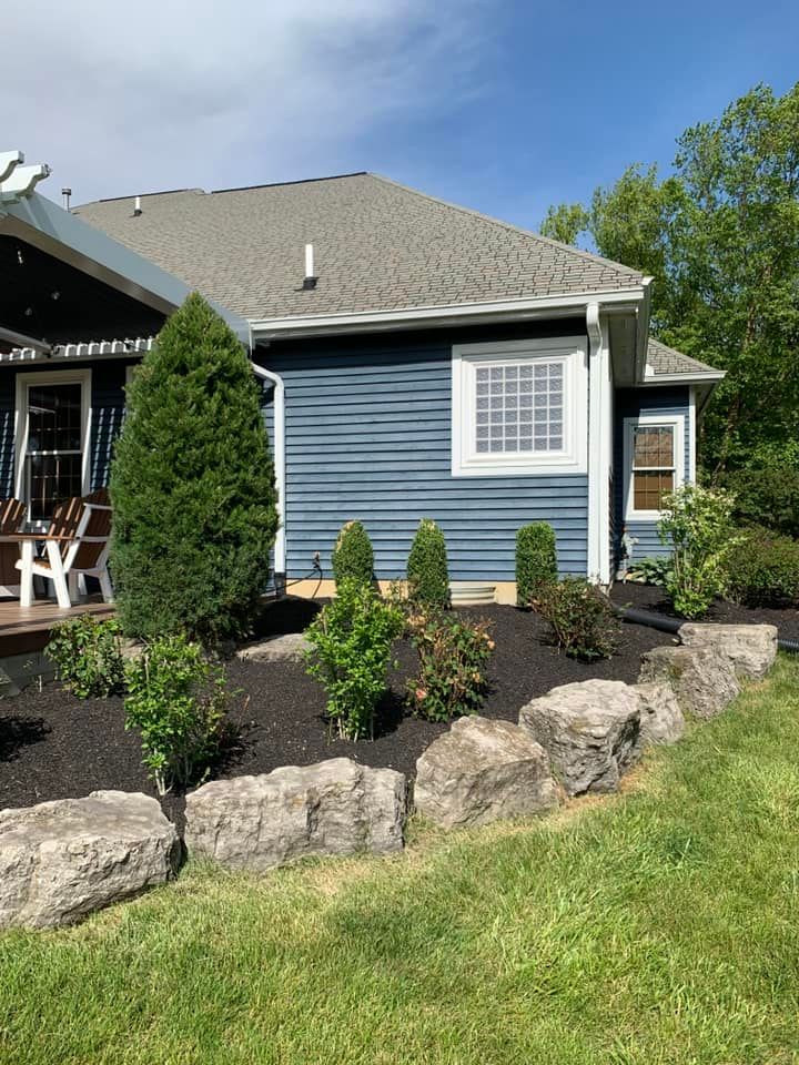 Blue house with dark mulch flower bed, bordered by rocks and green lawn.
