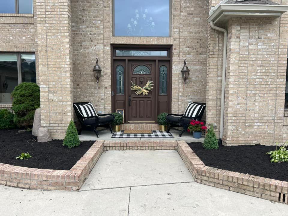 A brick home's front entrance features a dark door, two chairs, and a welcome mat.