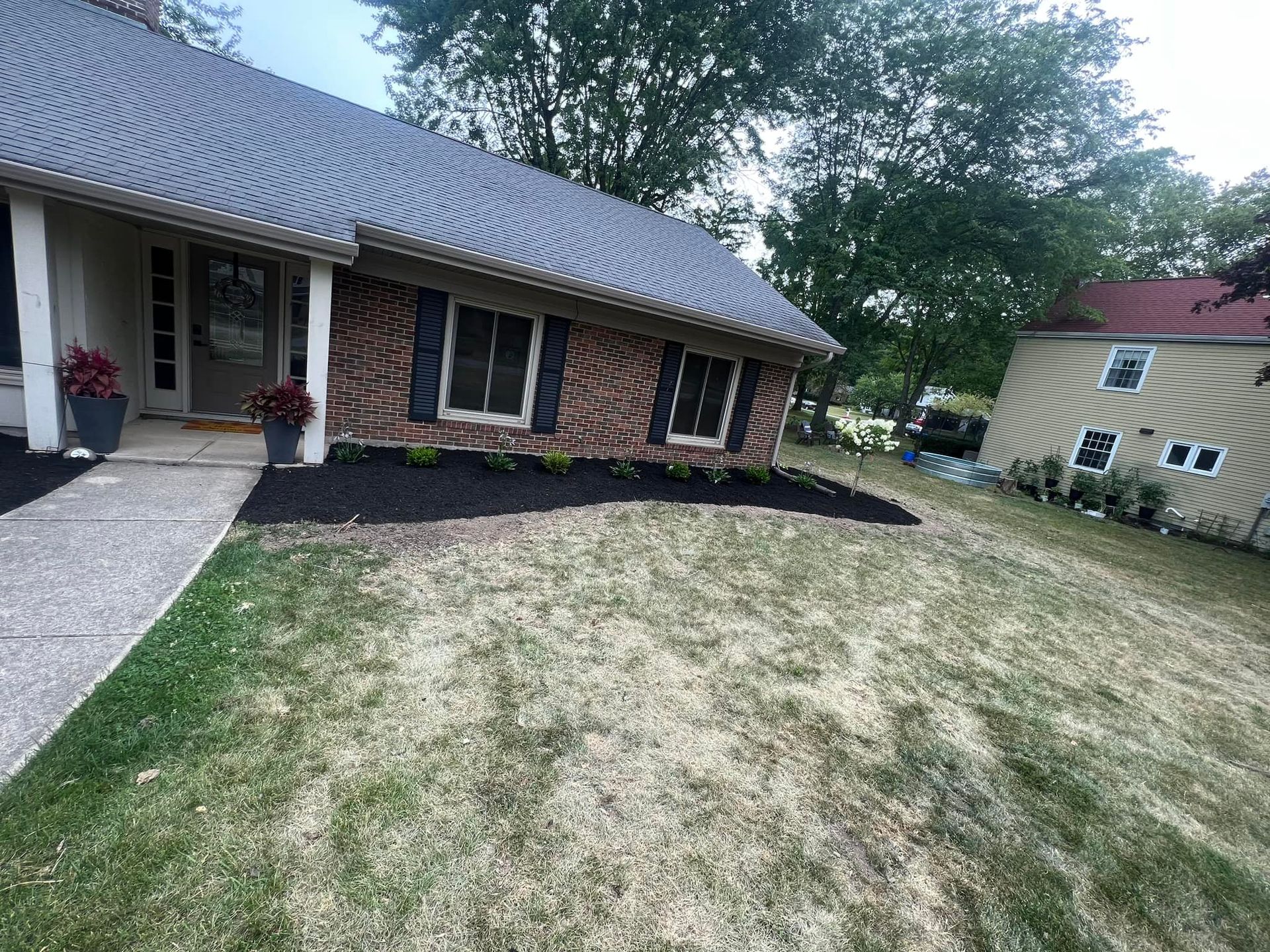 Brick house with black shutters, dark mulch border, green grass, and a smaller house on the right.