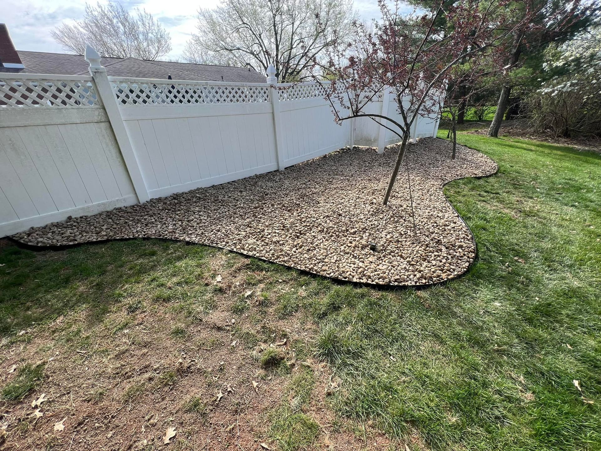 A decorative rock bed lines a white fence and small tree, edged with black, in a yard with patchy grass.