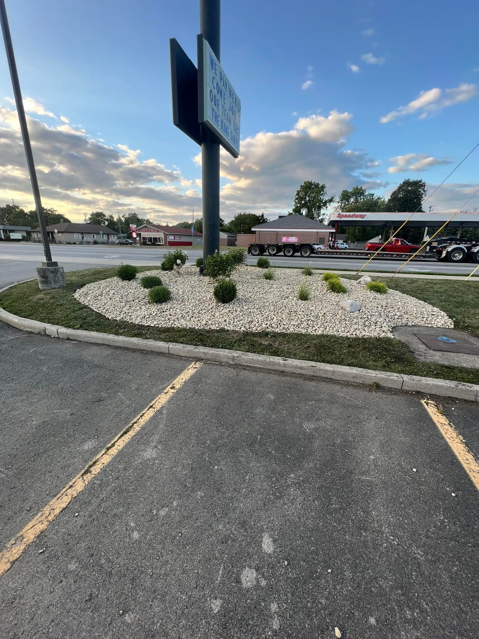 Flower bed in parking lot with sign post, rocks, and green plants. Semi-trucks in background.