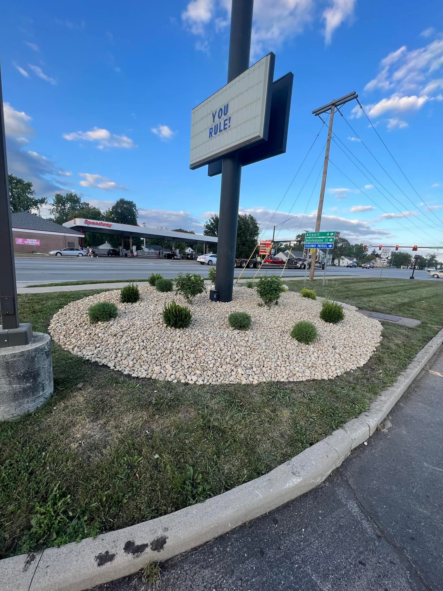 Round landscaping bed with bushes and light-colored stones around a signpost in front of a gas station on a sunny day.