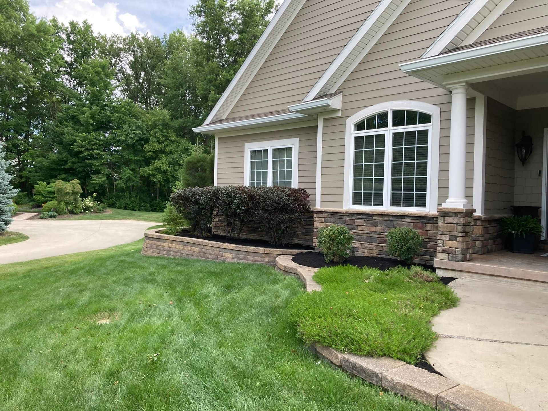 Tan house with stone facade and lush green lawn; flowerbed with dark bushes in front.