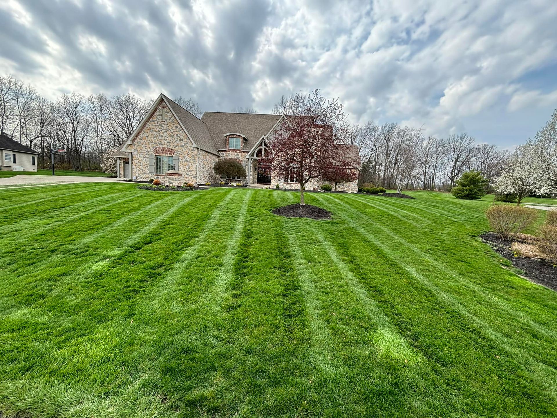 Lawn with neat mowing stripes in front of a stone house, under a cloudy sky.