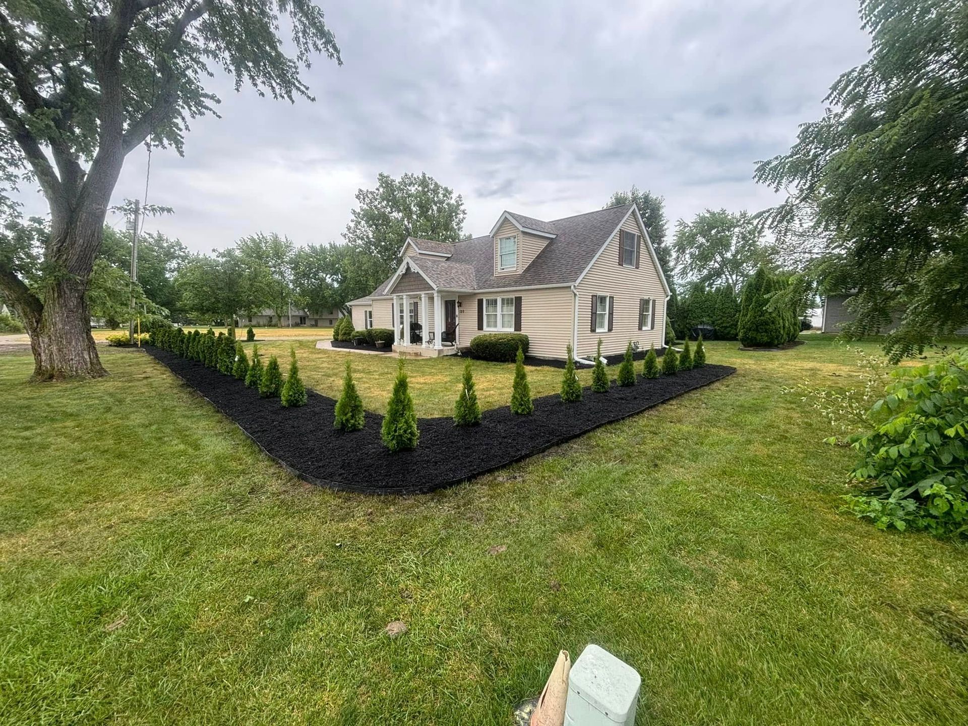 Curb appeal of a tan house with dormers, fronted by a black mulch bed and evergreen trees on a green lawn.