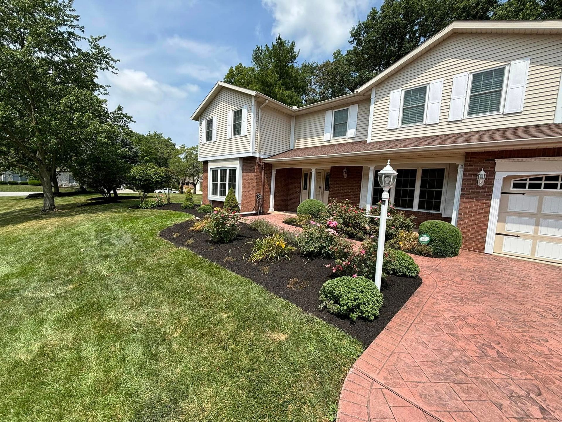 Two-story house with brick accents, dark mulch flowerbed, and a red-stamped driveway on a sunny day.