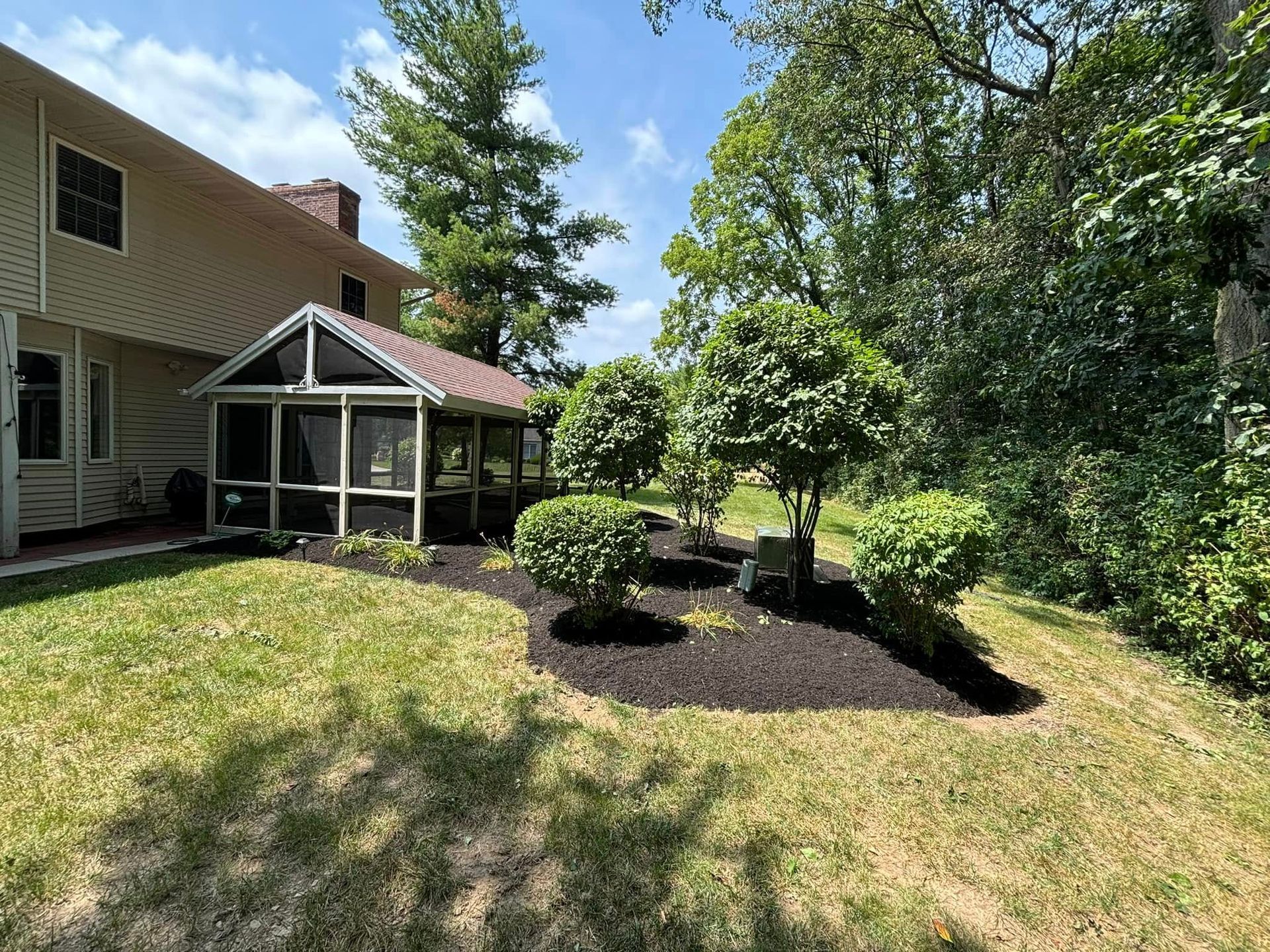 A screened porch next to a beige house; landscaping features bushes and mulch in a sunny yard.
