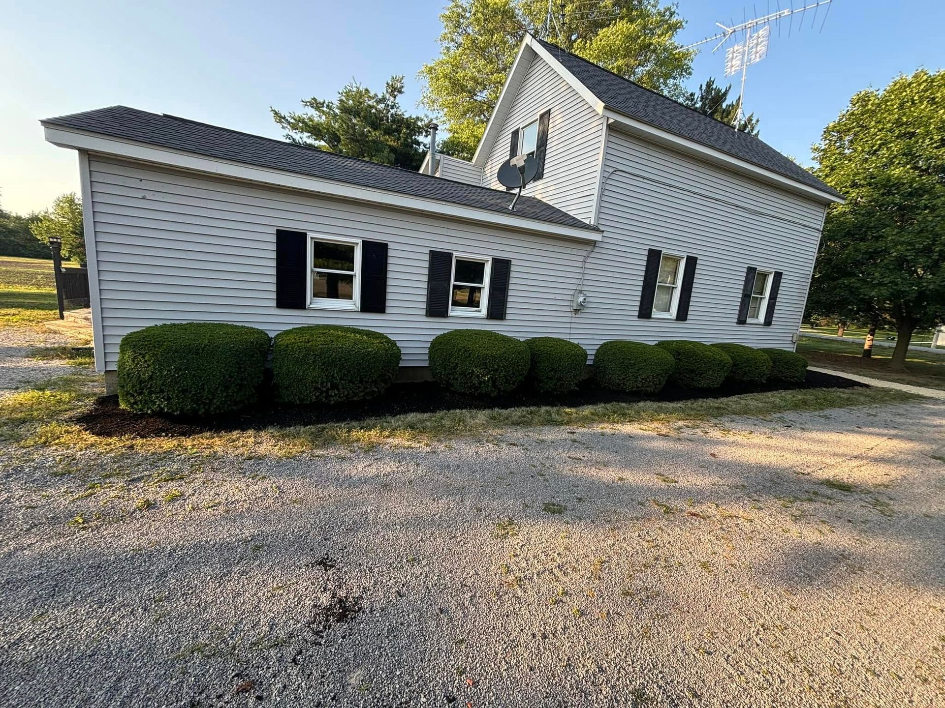 An angled house sculpture with black shutters, surrounded by green bushes and gravel, set outdoors.