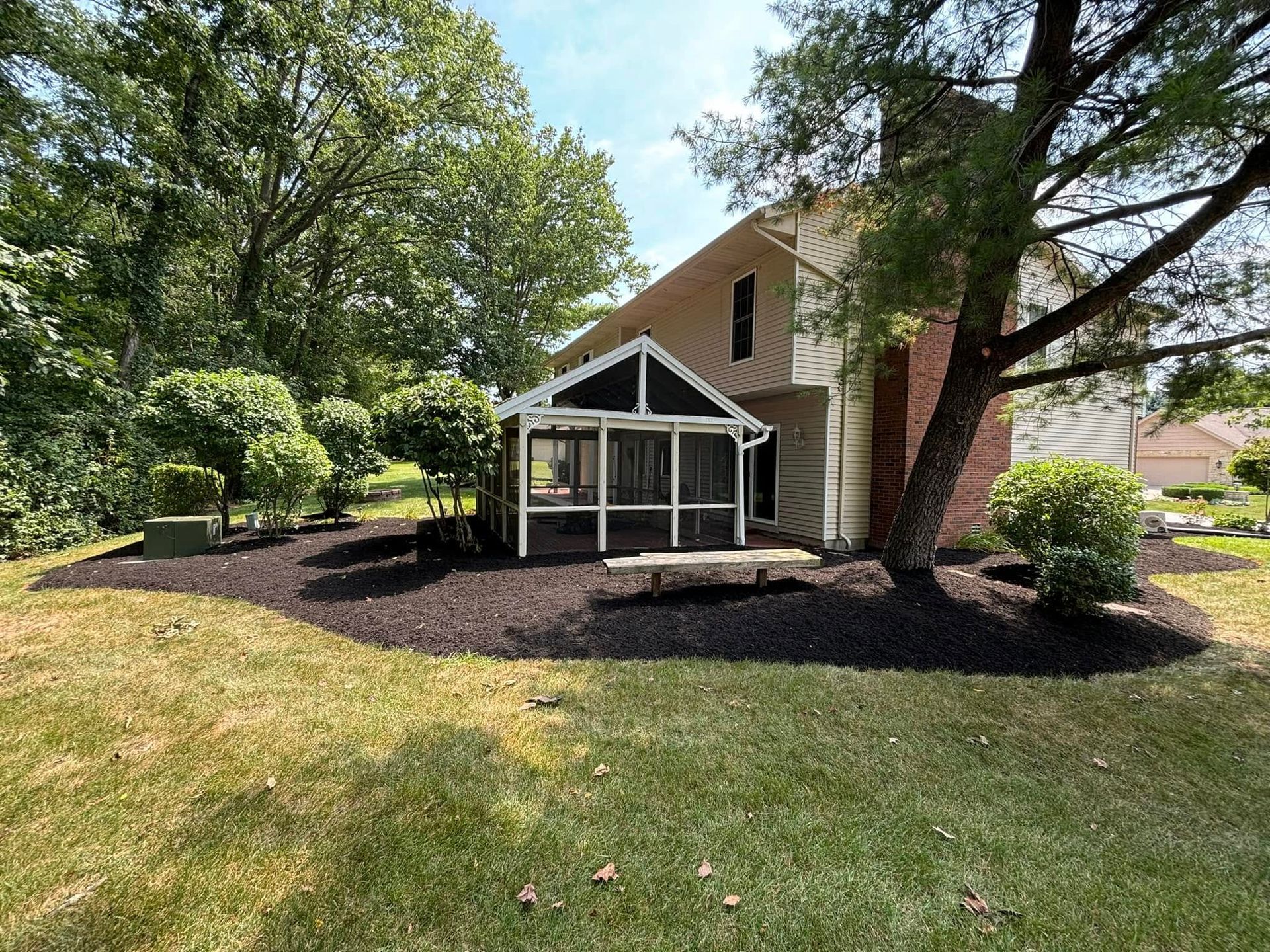 House exterior with a screened porch, surrounded by mulch and a lawn, under a sunny sky.
