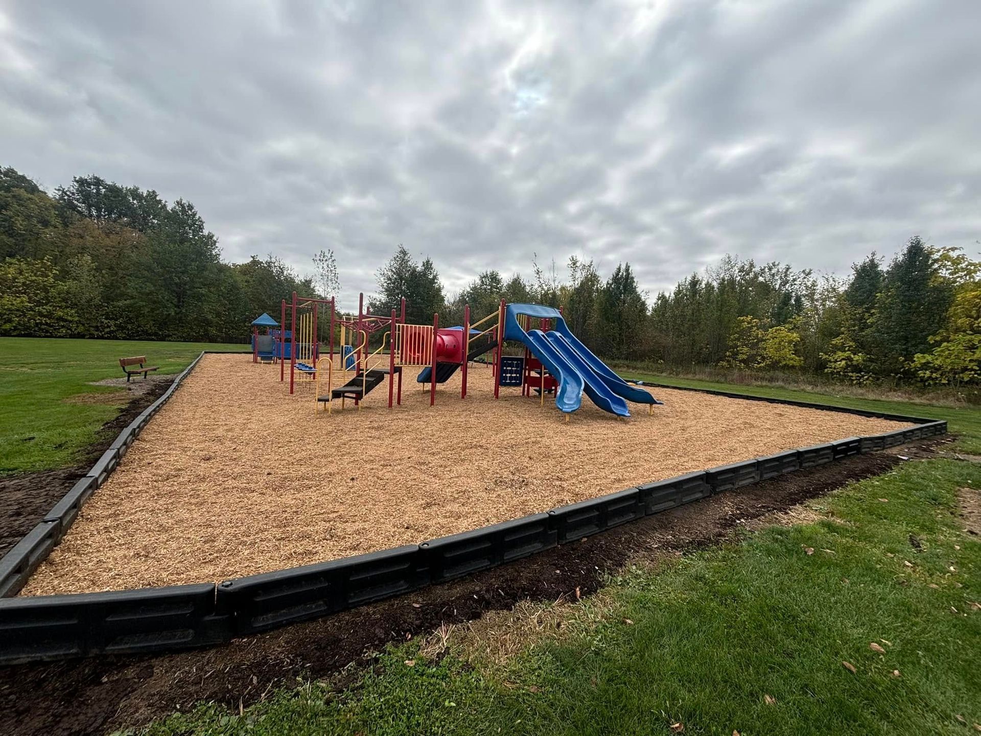 Playground with colorful equipment on wood chips, bordered by black edging, under a cloudy sky.