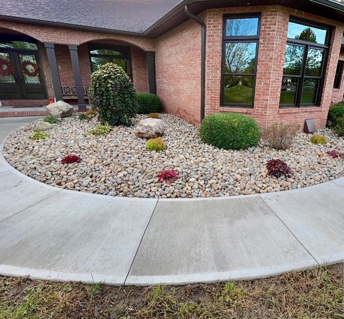 Circular flower bed with large rocks, shrubs, and red plants in front of a brick house with a curved sidewalk.
