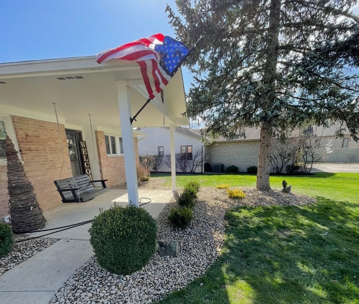 A house with an American flag hanging from the porch. There's a porch swing and neatly trimmed shrubs in the yard.