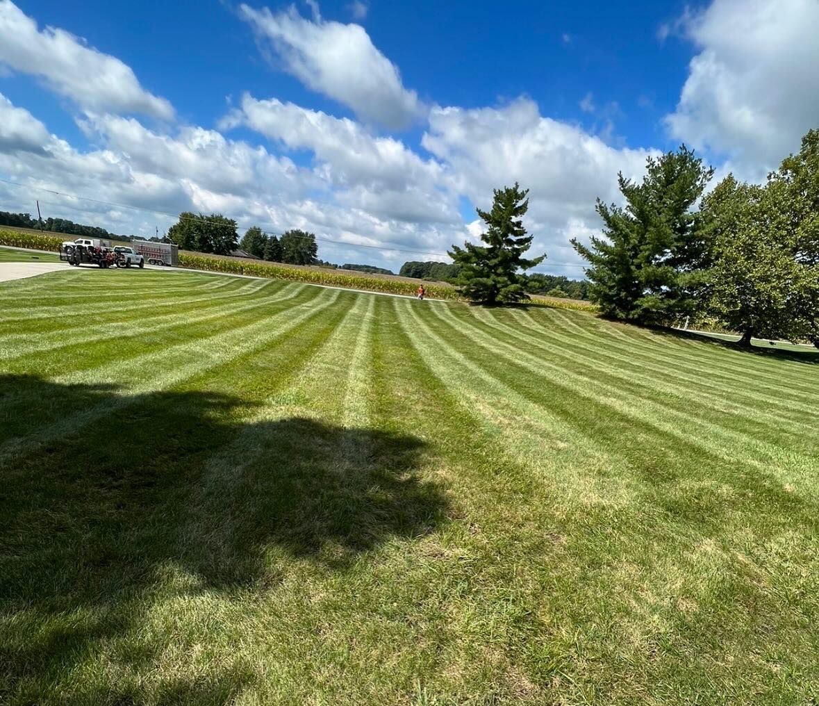 Lawn mowed in stripes under a blue sky with clouds and trees.