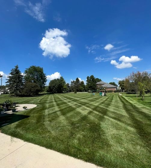 Lawn with a diamond-patterned mowing design under a blue sky with clouds. Trees and a house are in the background.