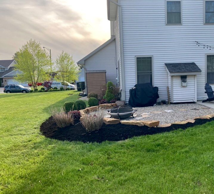 Backyard patio with a fire pit, surrounded by landscaping and a grassy lawn. House in the background.