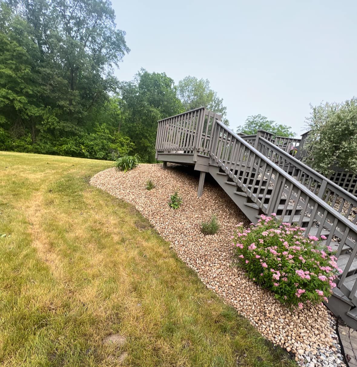 Wooden deck with stairs, surrounded by a grassy lawn and gravel garden bed with flowering shrubs.