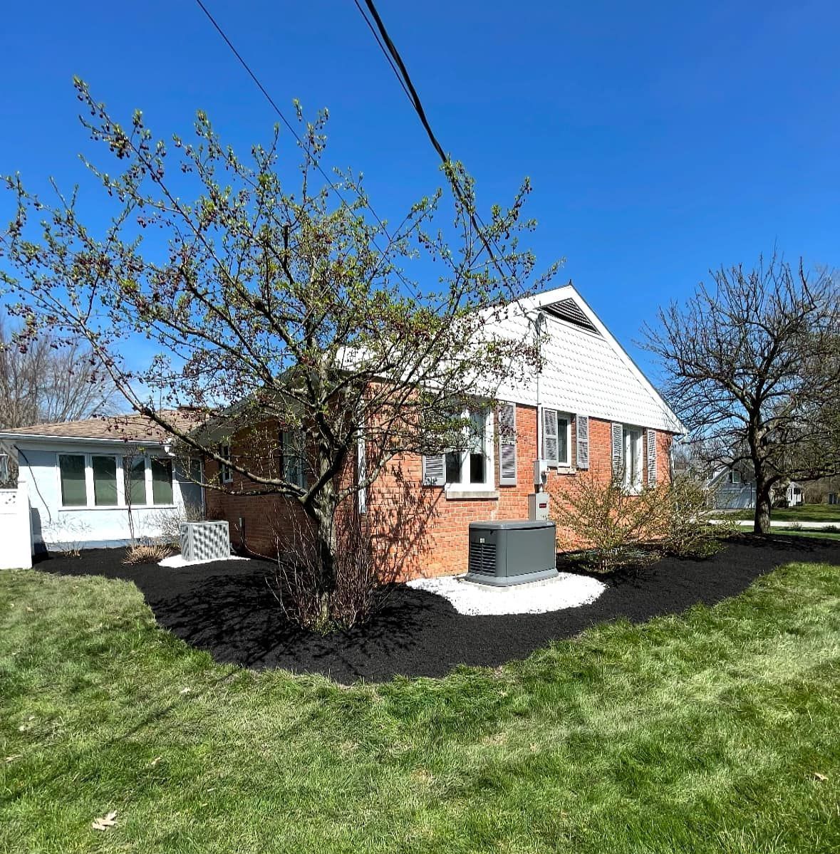Red brick house with a white roof and a lush green lawn on a sunny day. A tree partially obscures the front of the house.