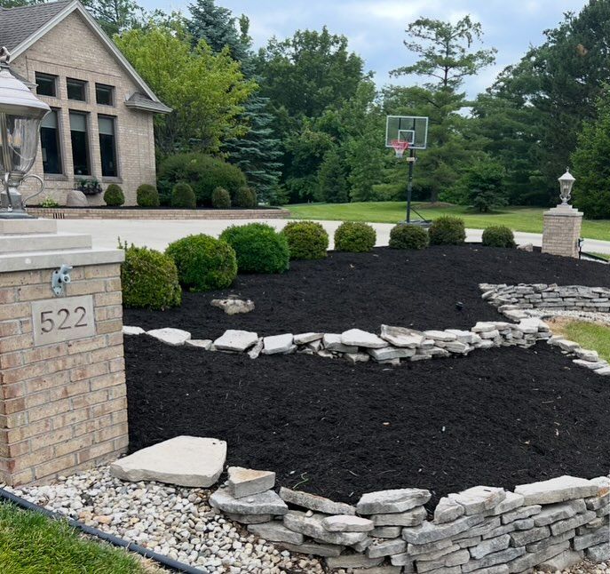 Brick house with black mulch landscaping, stone border, green bushes. Basketball hoop in the background.