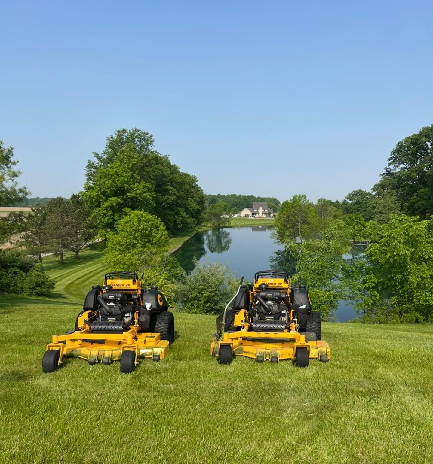 Two yellow lawn mowers on a grassy hill overlooking a pond and a house on a sunny day.