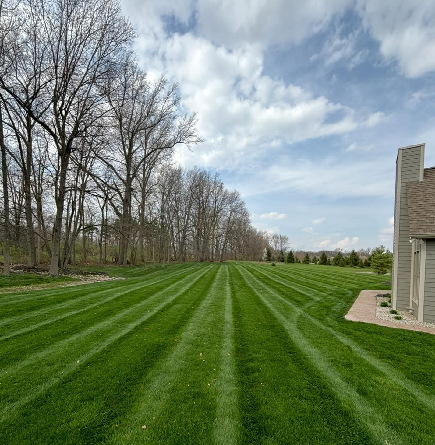 Green lawn with striped mowing pattern, trees on the left, a house on the right, and a cloudy sky.