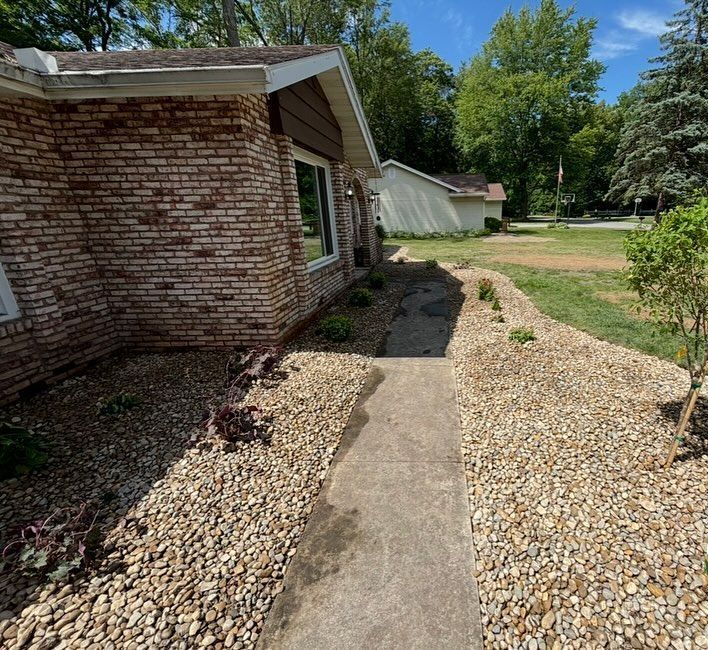 Brick house with a concrete path, bordered by gravel and small plants, leading to a distant building.