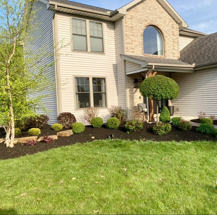 Two-story house with beige siding and brick facade. Landscaped yard with black mulch and various green plants.