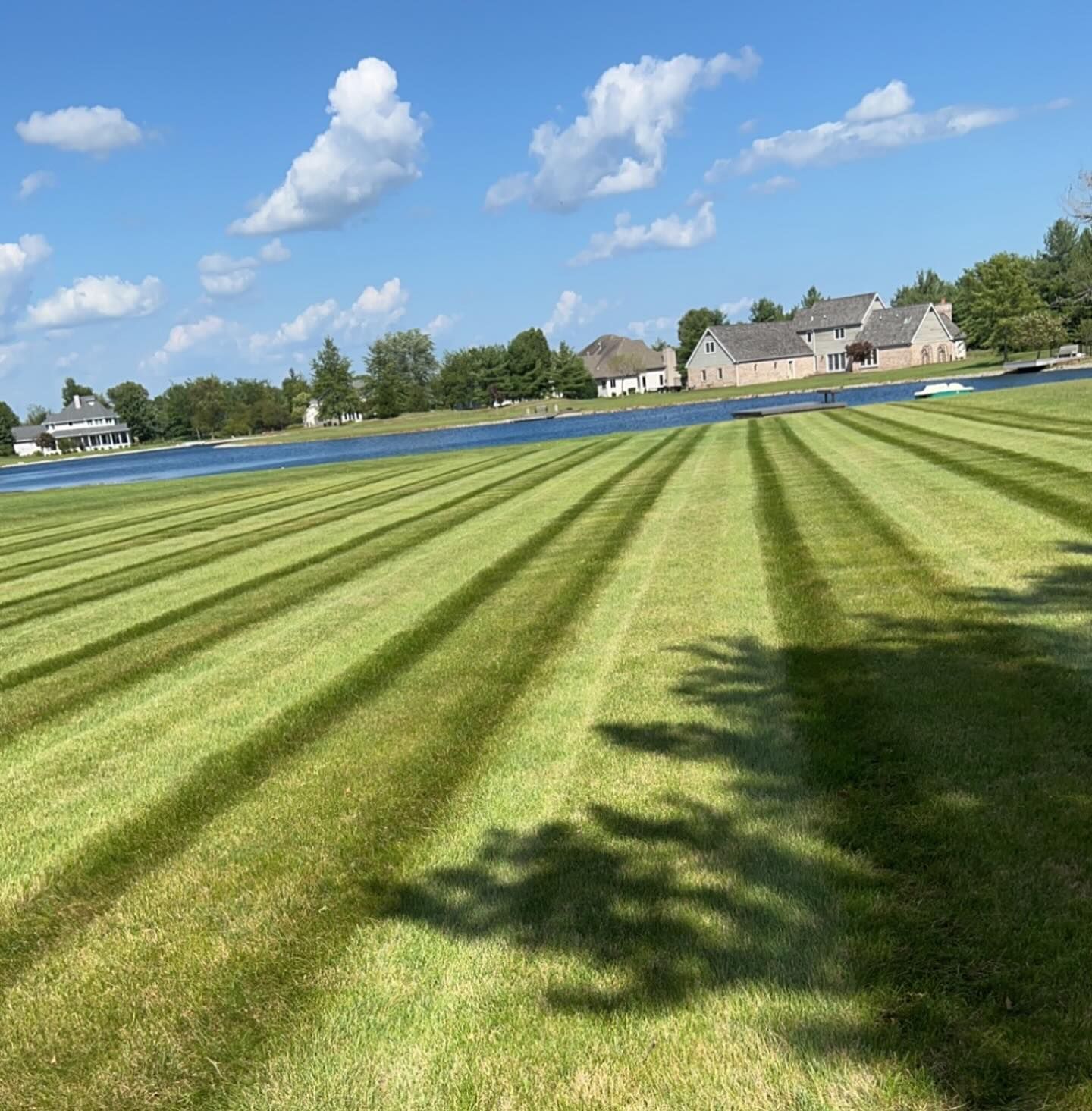 Lawn mowed in striped patterns near a lake with houses and a blue sky with clouds in the background.