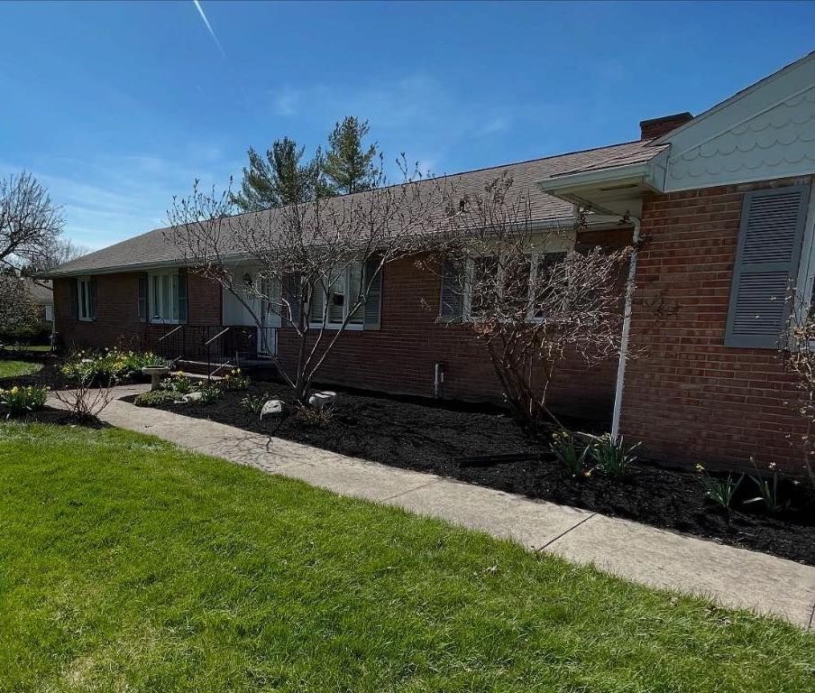 Brick ranch house with a pathway leading to the front door, with greenery and trees in the yard.
