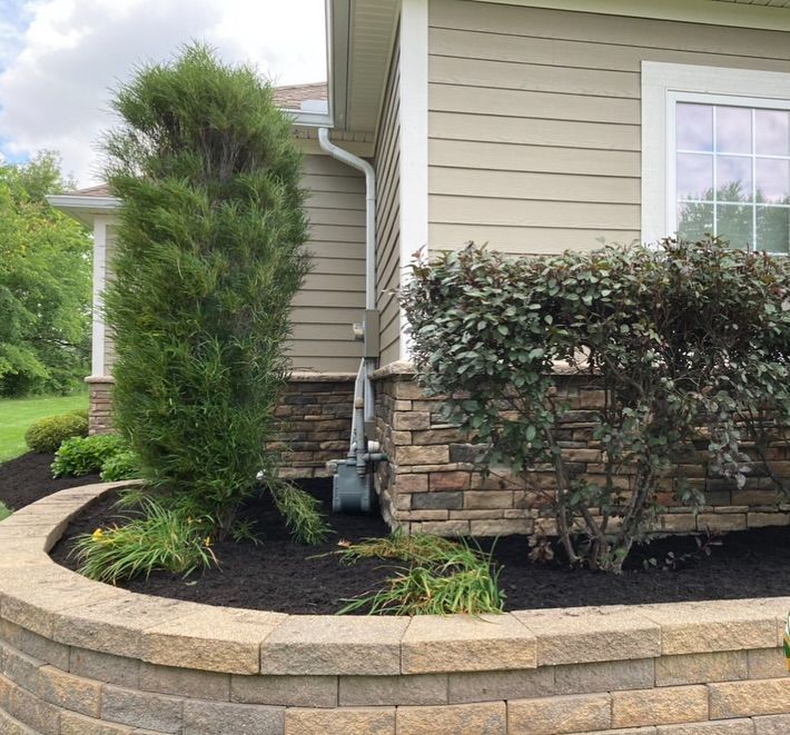 A house corner with tan siding, stone accents, and a curved retaining wall with dark mulch and shrubbery.