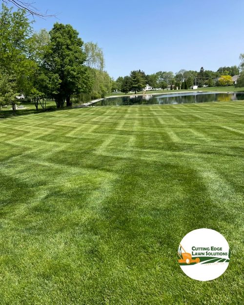 Green lawn with geometric mowing patterns, a pond in the background, and a sunny sky. Logo in the bottom right corner.