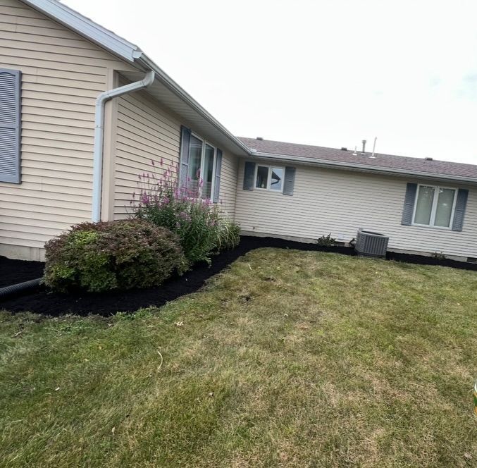 House with tan siding, grey shutters, and black mulch in flower bed. Green grass lawn.