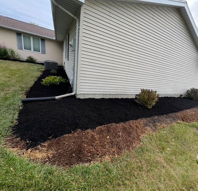 Black mulch borders a house with white siding, plants, and a downspout.