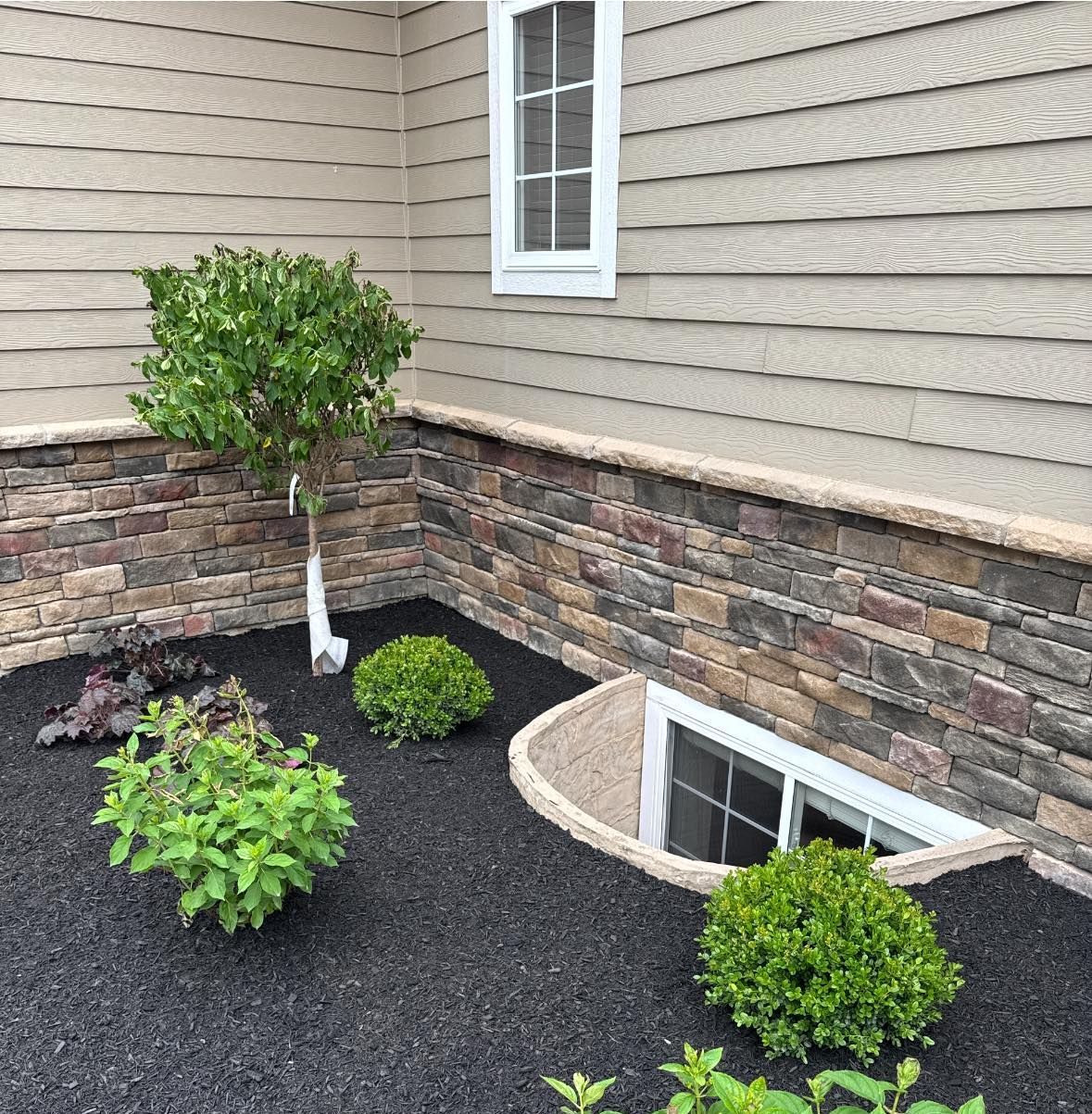 Corner of a house with a stone-covered foundation and a small garden bed filled with plants and dark mulch.