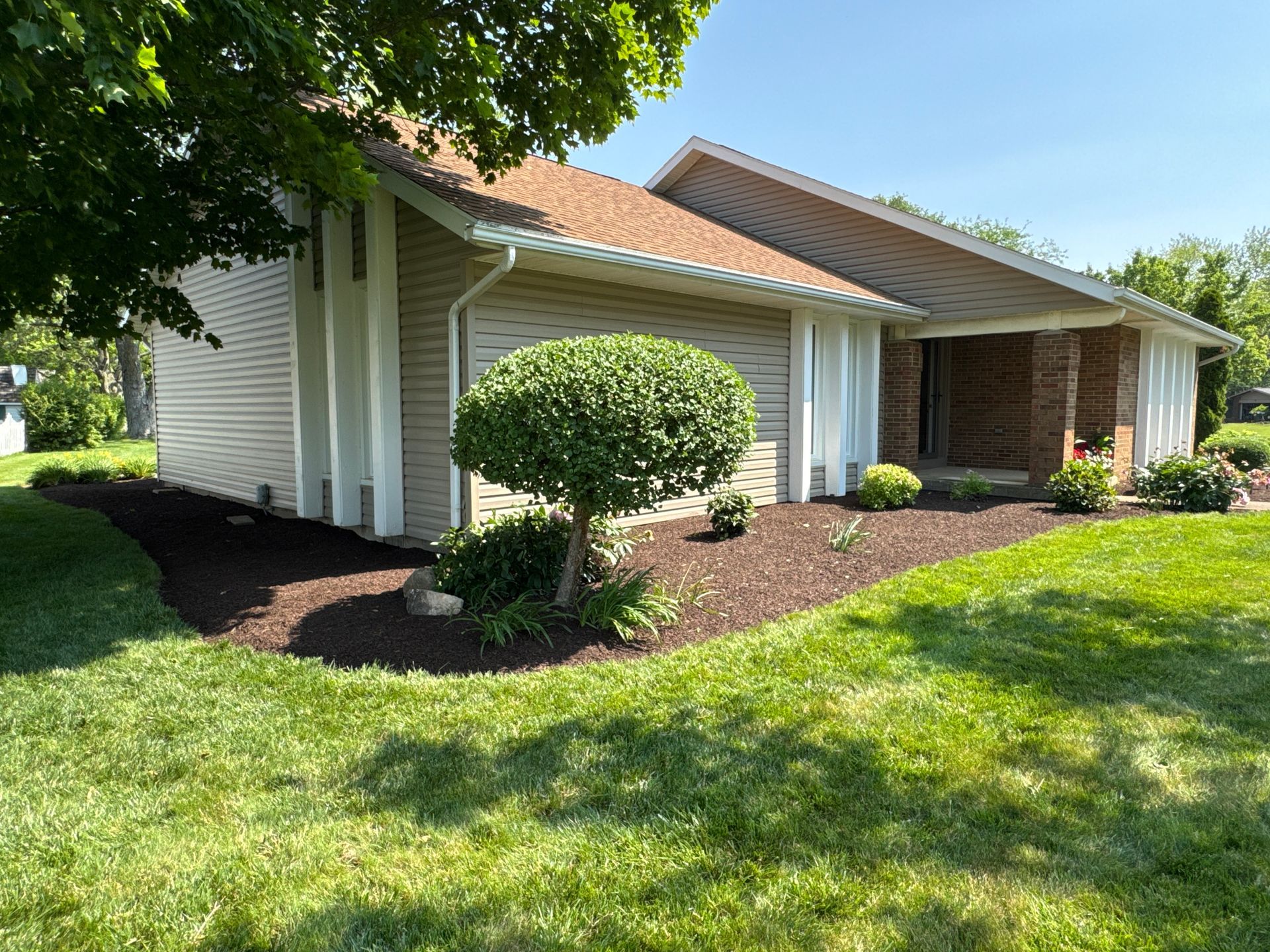 Beige house with brown roof, lush green lawn, and flower bed with dark mulch.
