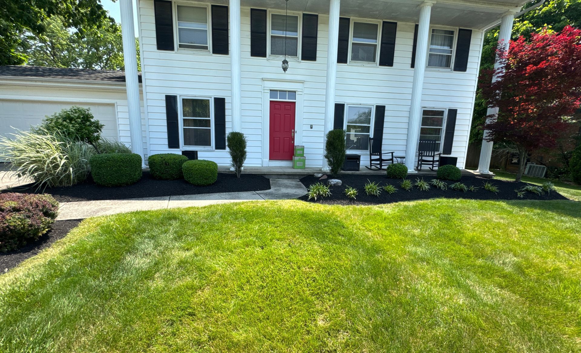 Two-story white house with black shutters, red door, lush green lawn, and well-maintained landscaping.