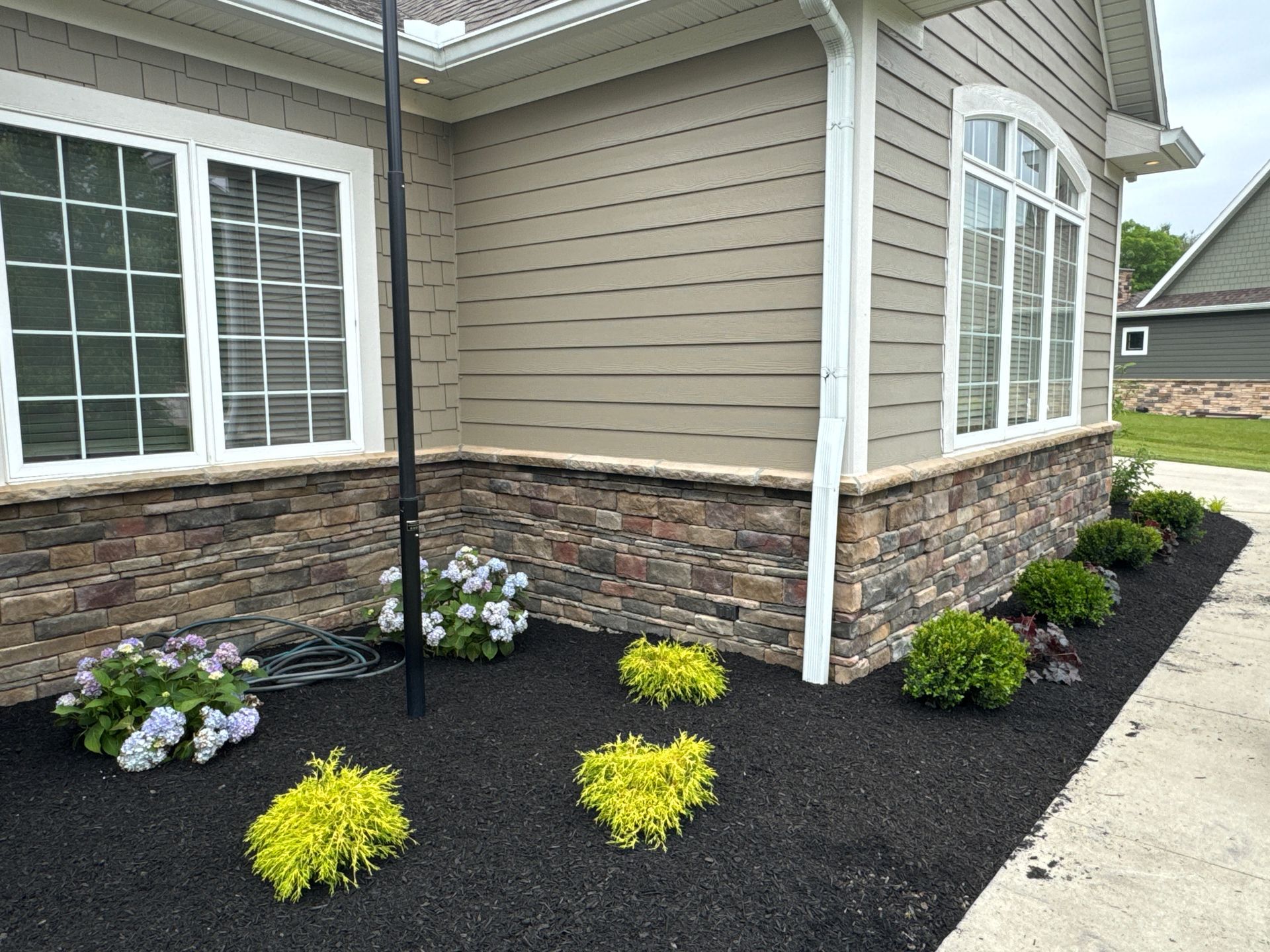 House exterior with stone veneer, tan siding, white windows, and a flower bed with black mulch and green and yellow plants.