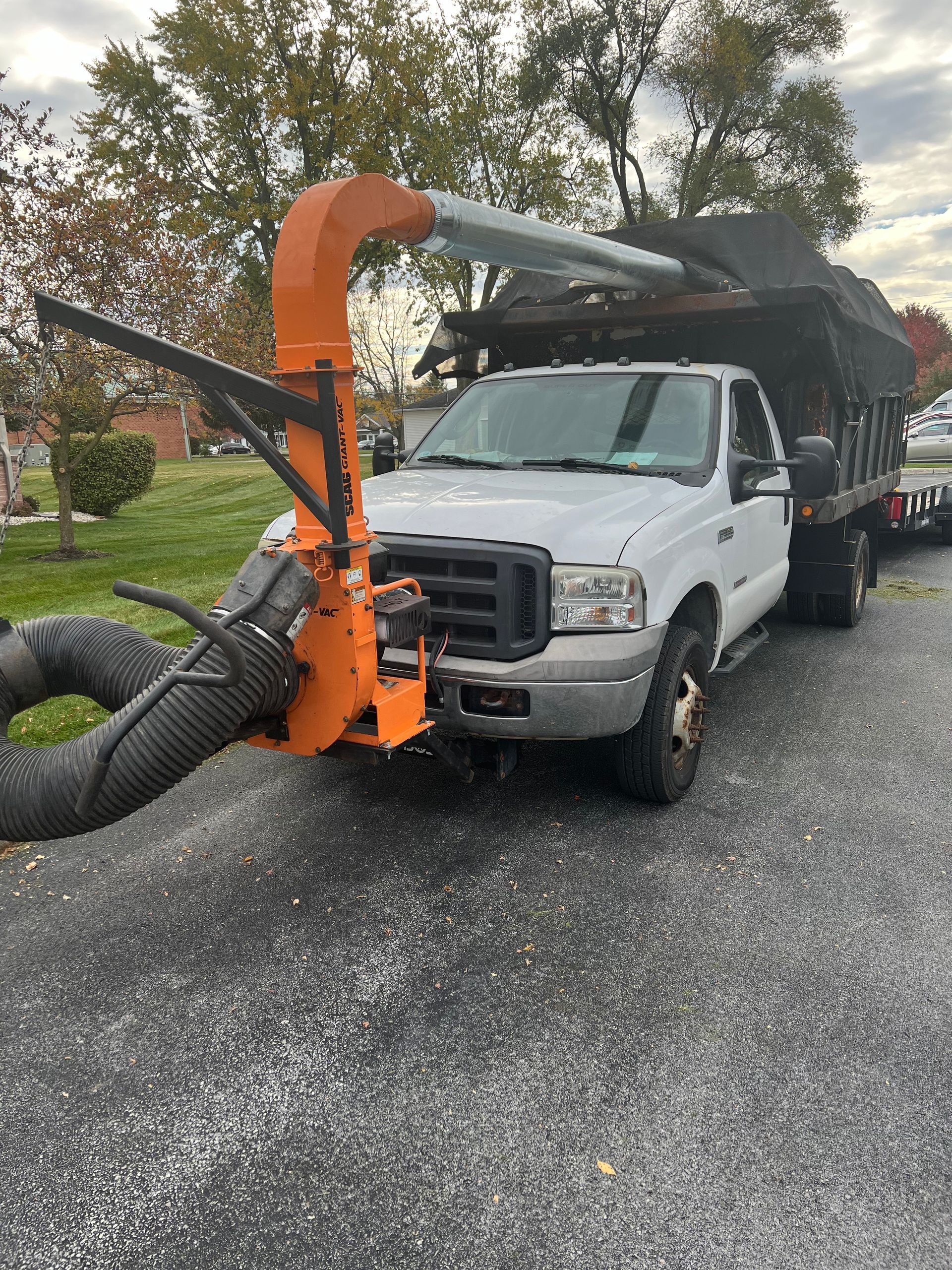 White pickup truck with an orange leaf vacuum attached, parked on a driveway; trees and grass in background.