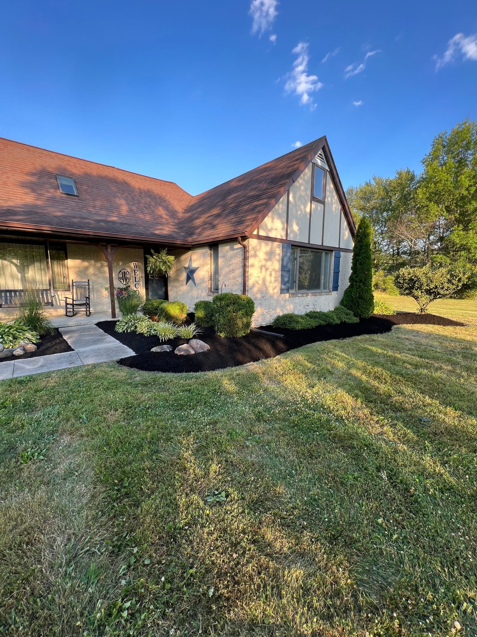 Tudor-style home with brown roof, tan stucco, and dark mulch. Lush green lawn with blue sky background.