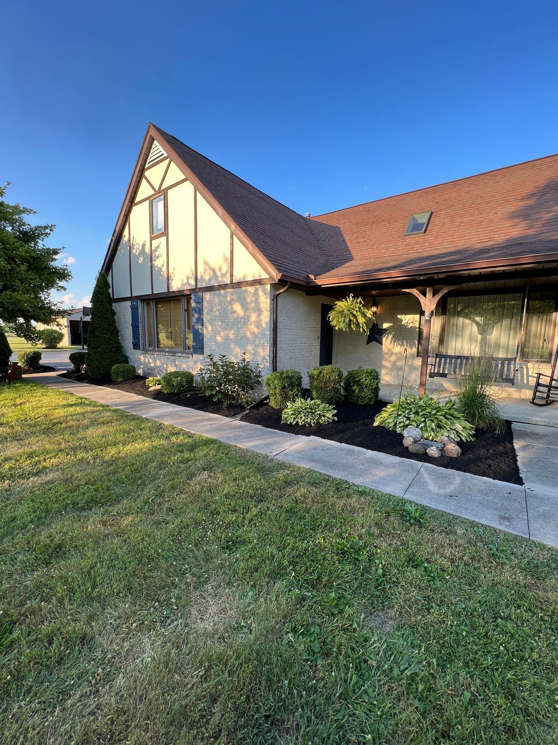 Tudor-style house with brown roof, beige and tan walls, and manicured landscaping. Clear blue sky in the background.