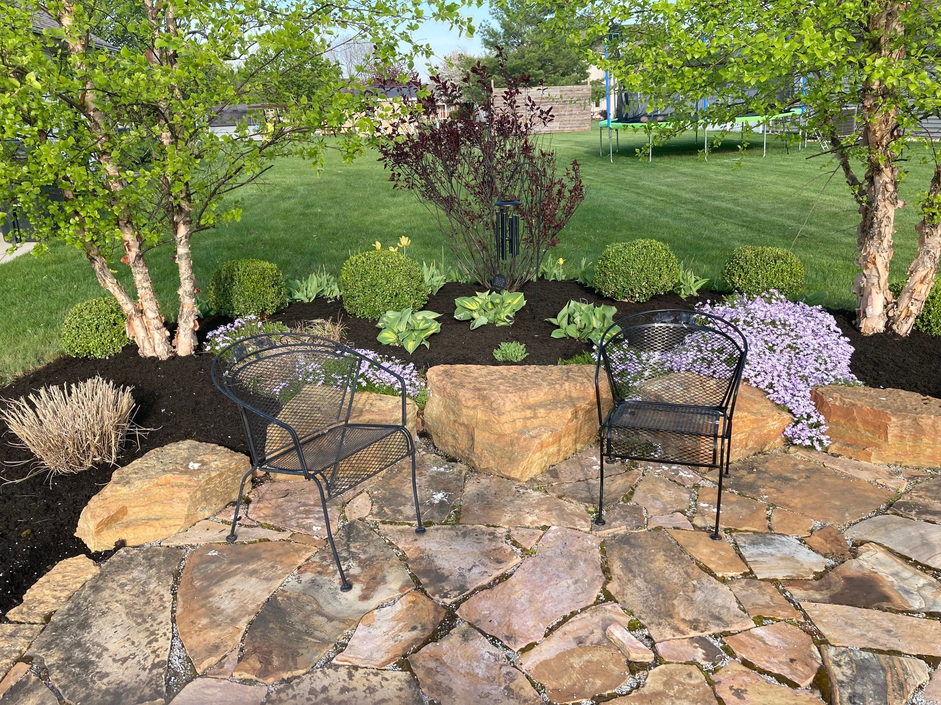 Stone patio with two black chairs, surrounded by landscaping and trees.