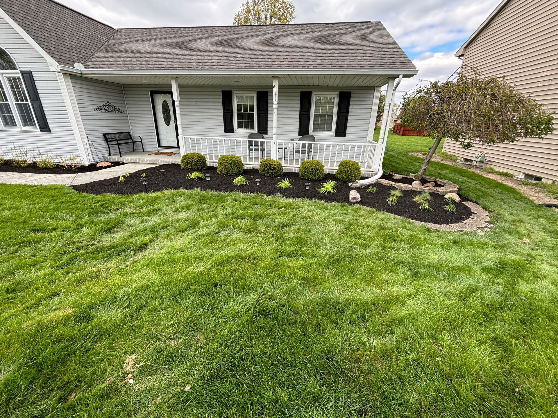 A light gray house with a porch, black shutters, and well-manicured lawn with fresh mulch and shrubs.