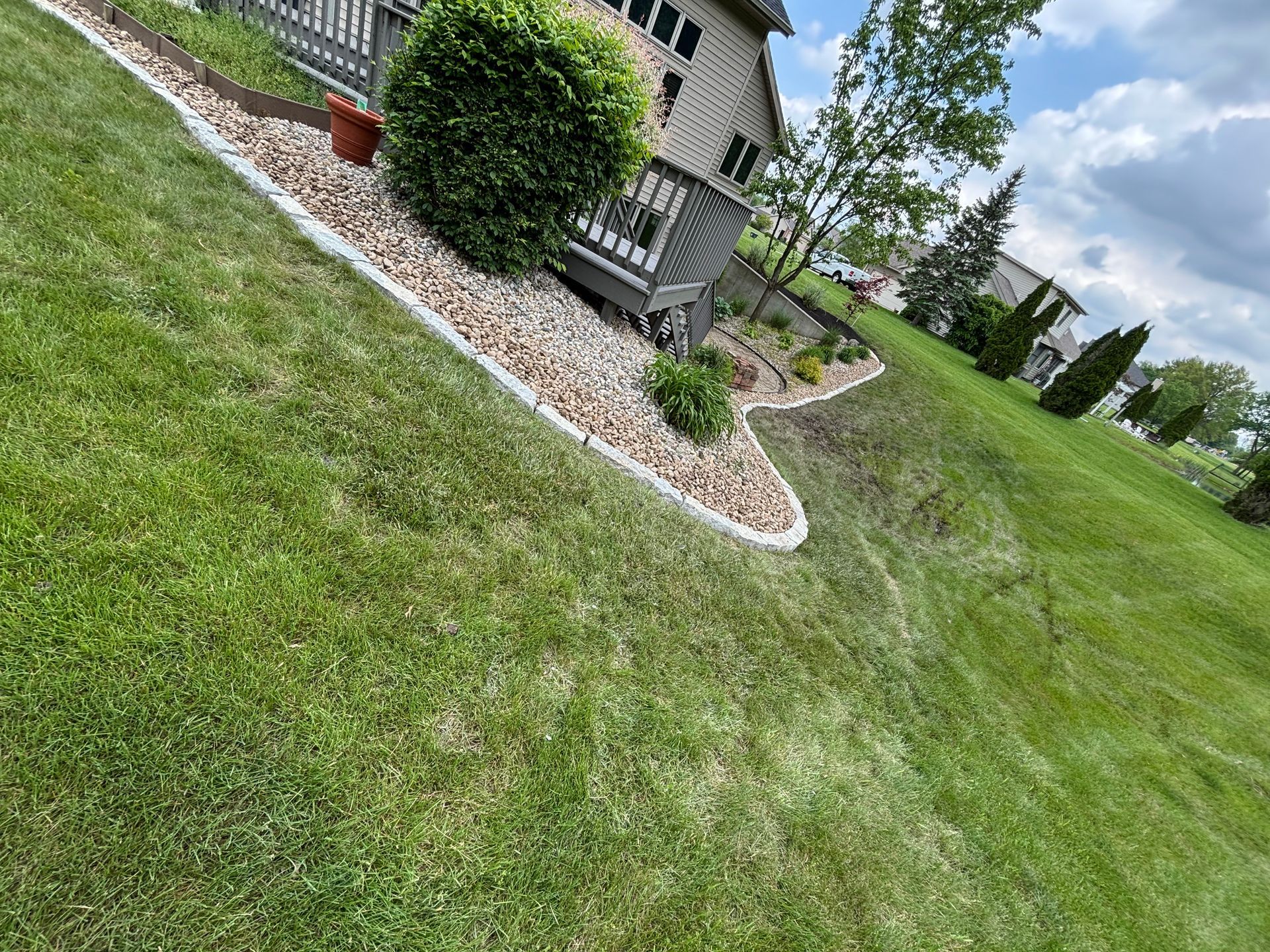 Lawn slopes down towards a house with a rocky landscape bed along the side. Blue sky with clouds.