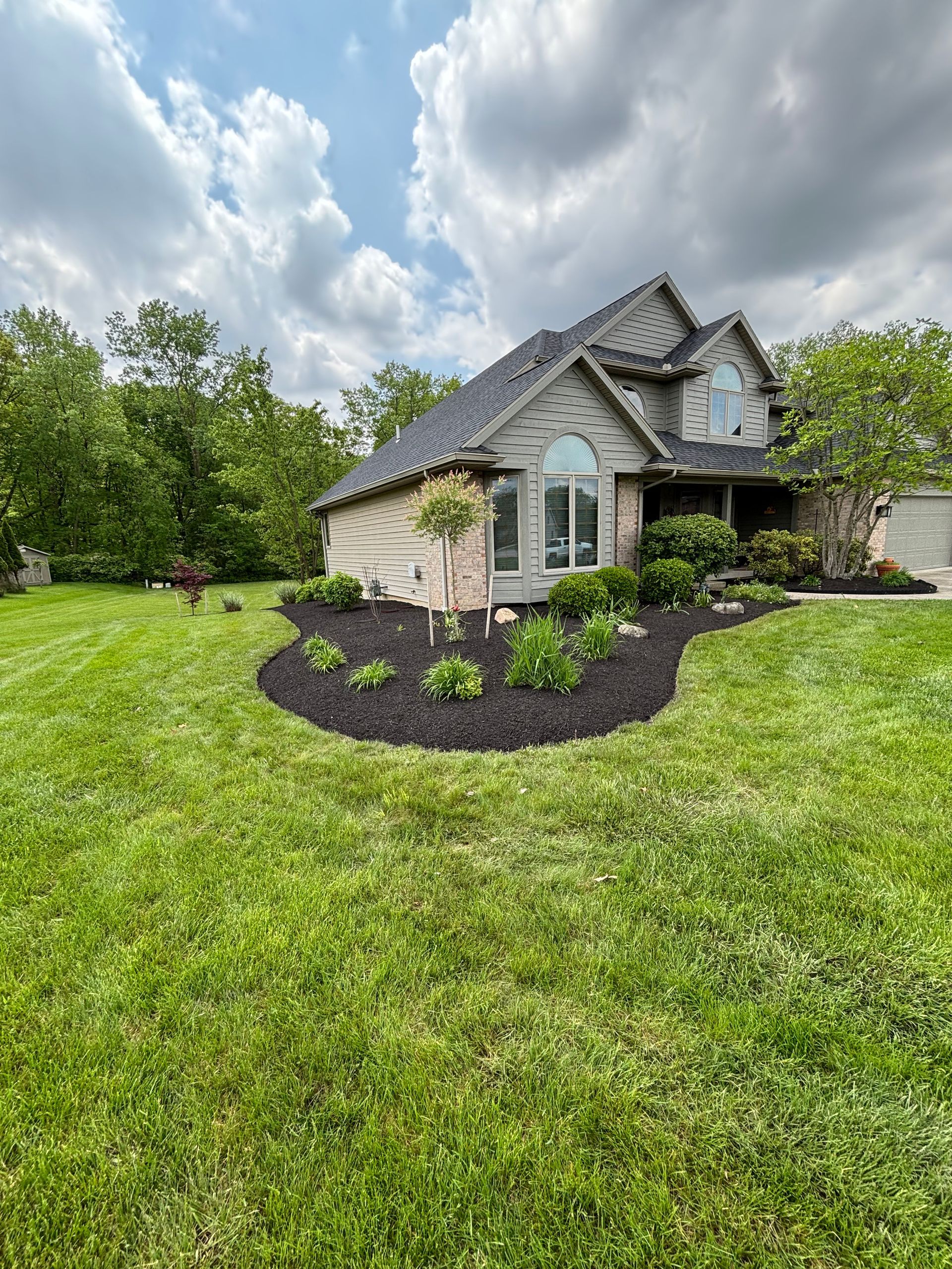 Two-story house with manicured lawn, black mulch flower beds, and cloudy sky.