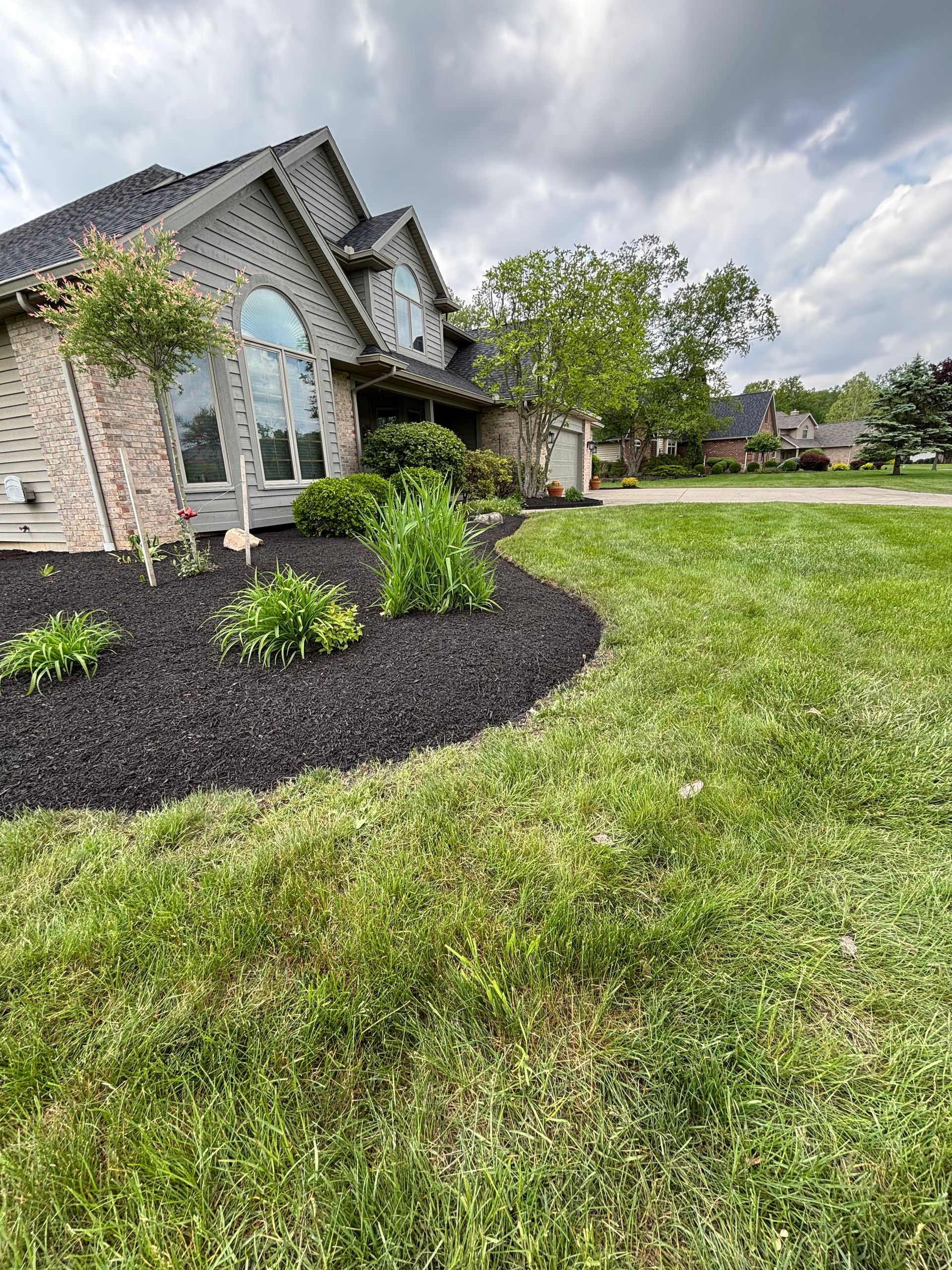 House with black mulch flower bed, green lawn, and cloudy sky.
