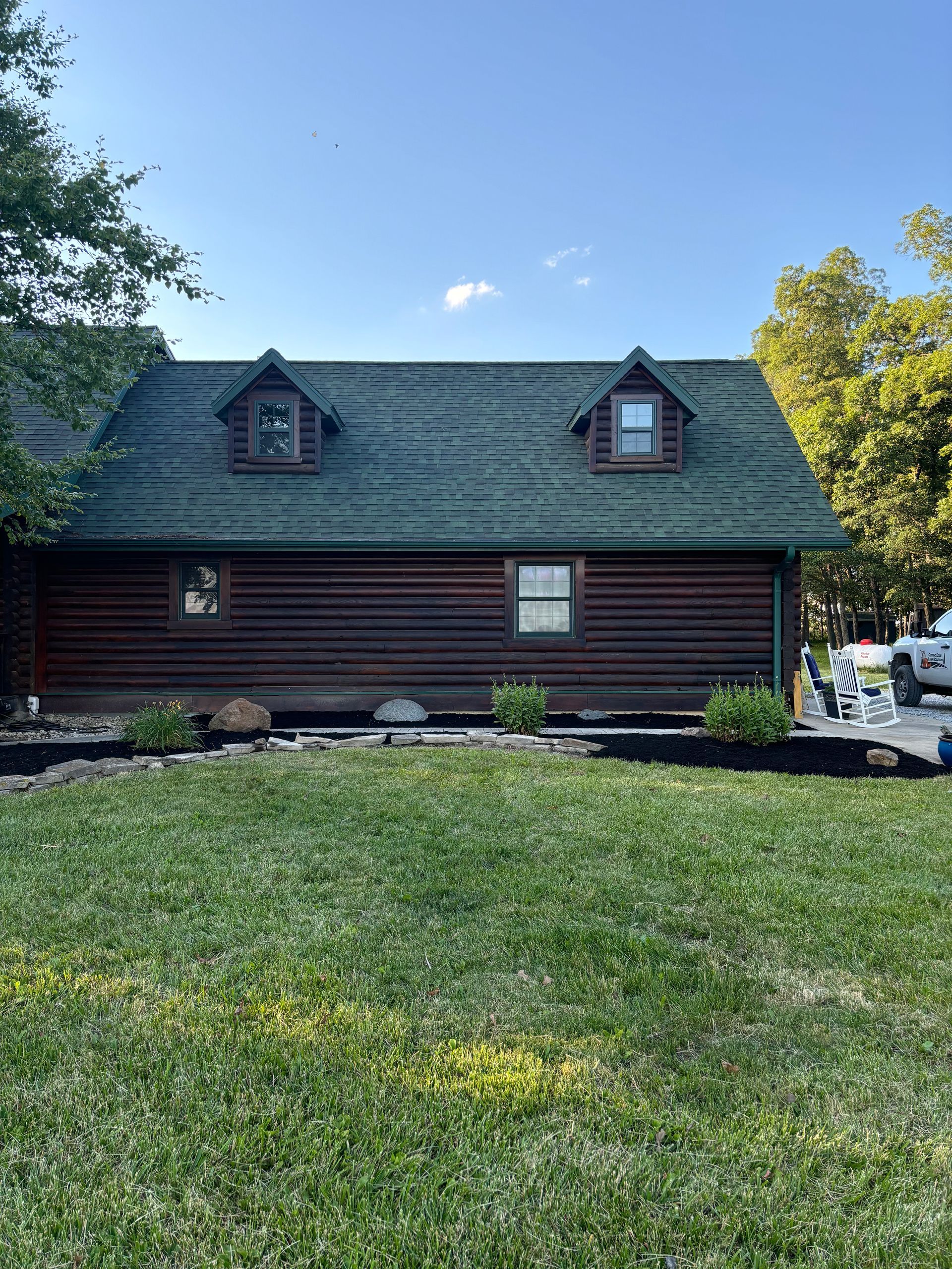 Log cabin with green roof and two dormers, surrounded by lawn and landscaping against a blue sky.