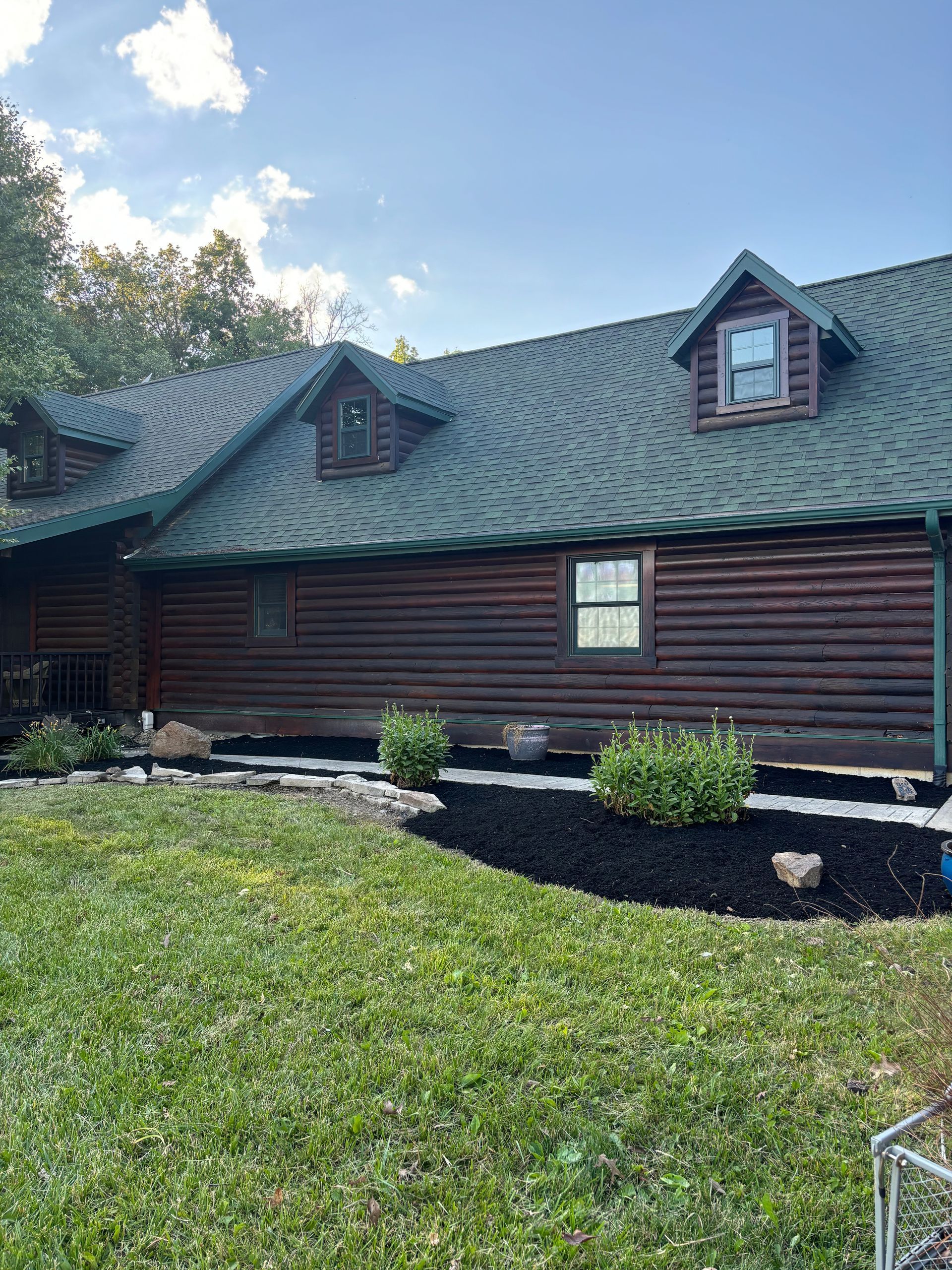 Log cabin with green roof, small dormer windows, and landscaping featuring fresh black mulch and green bushes.