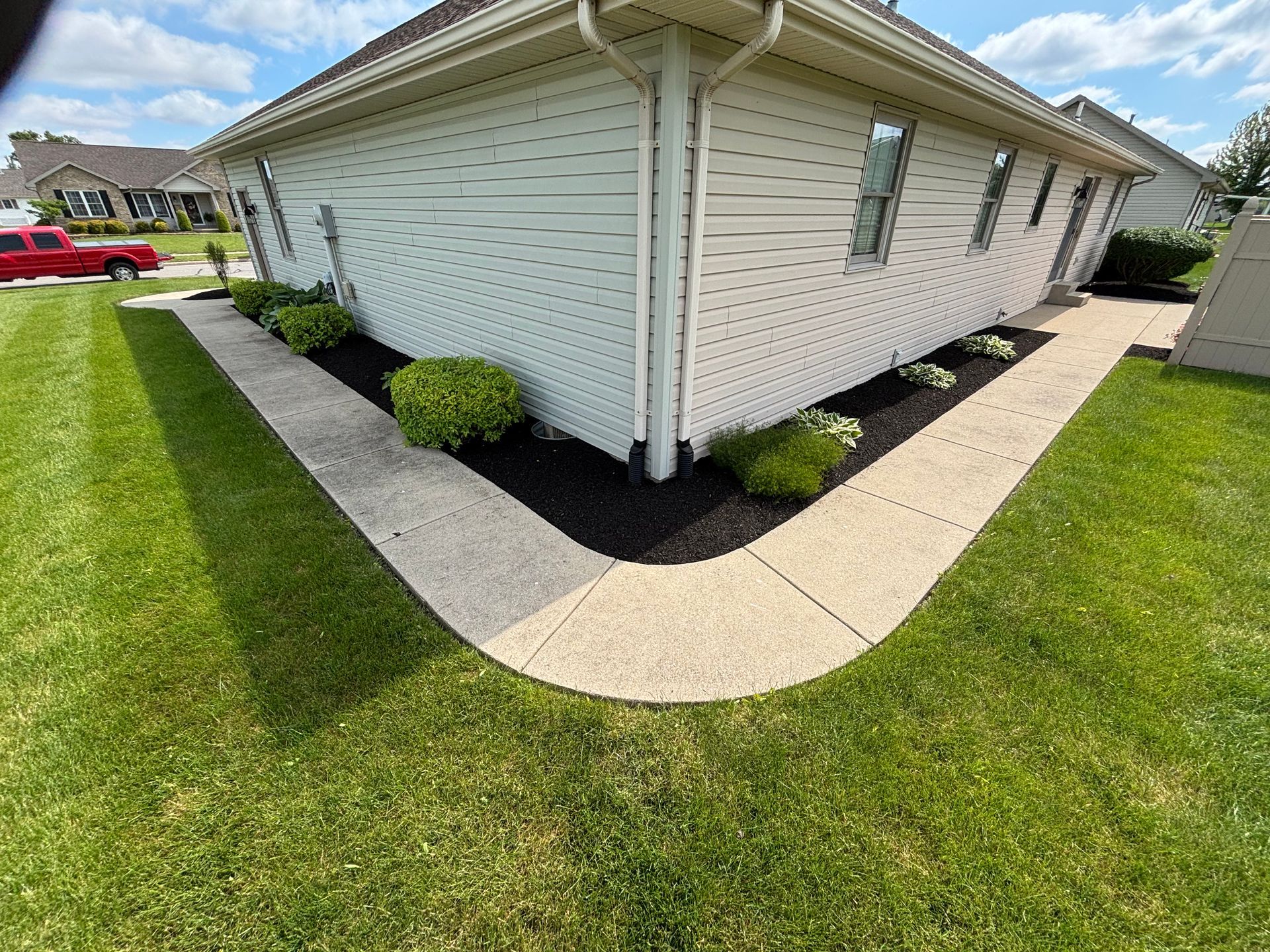 Corner of a house with a curved sidewalk, black mulch, green bushes, and grass.